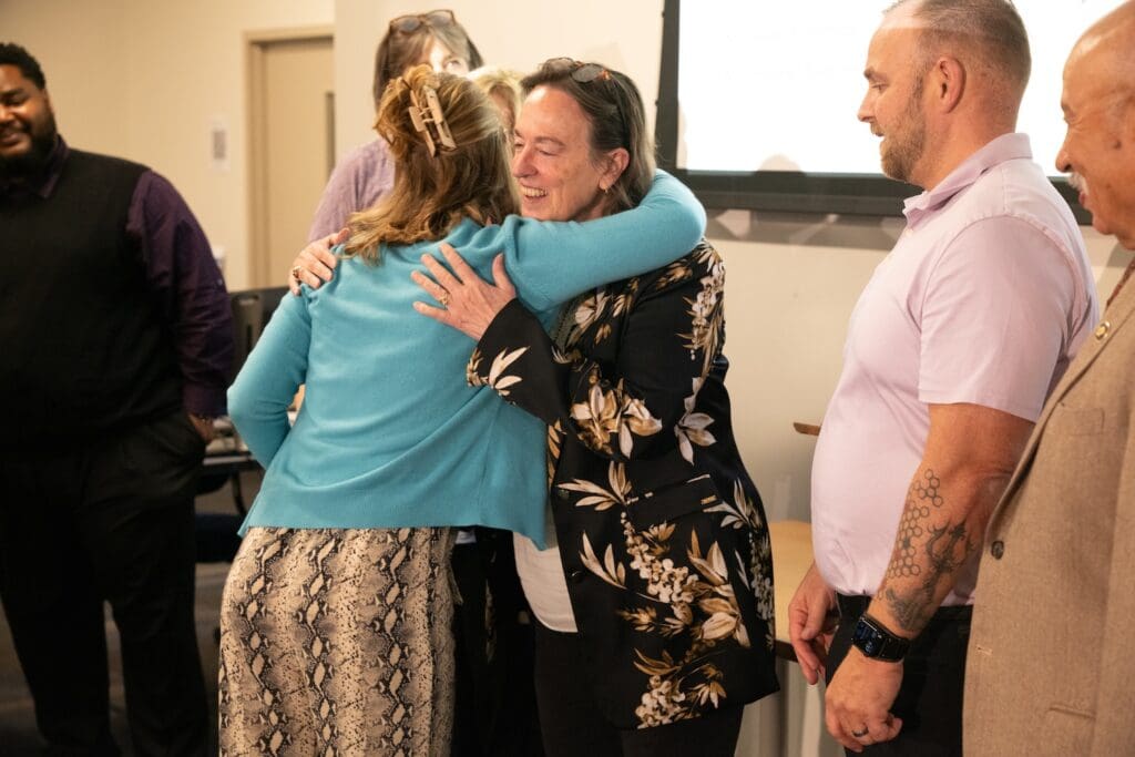 A group of Baltimore residents stands together, with one woman in a blue sweater warmly hugging another in a floral jacket. The scene reflects a moment of community engagement, as others nearby watch with the shared understanding and unity that strengthen race relations.