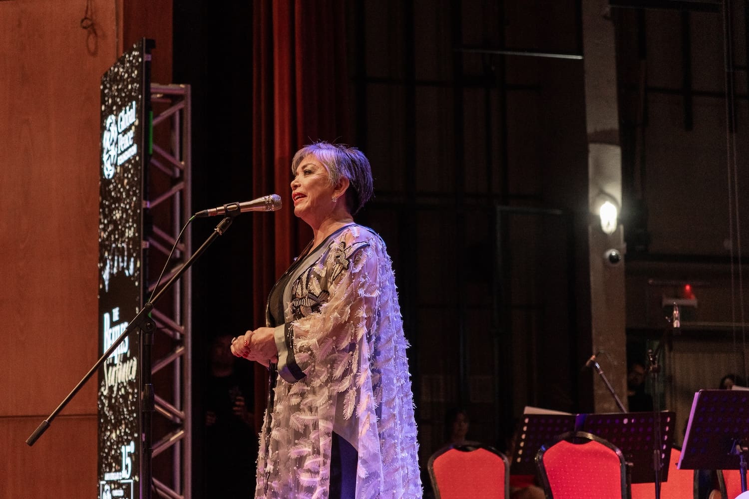 A person is speaking into a microphone on stage, wearing a patterned outfit that reflects their vibrant cultural heritage. Red chairs and a light display are visible in the background as they passionately discuss peacebuilding initiatives with the Global Peace Foundation in Paraguay.