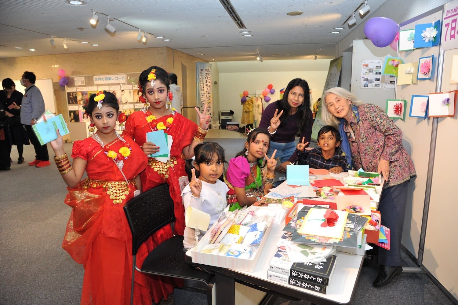 A multicultural group, including children in colorful attire, gathers around a table with crafts and art supplies inside a room adorned with festive displays and balloons, celebrating the Family Festival 2024.