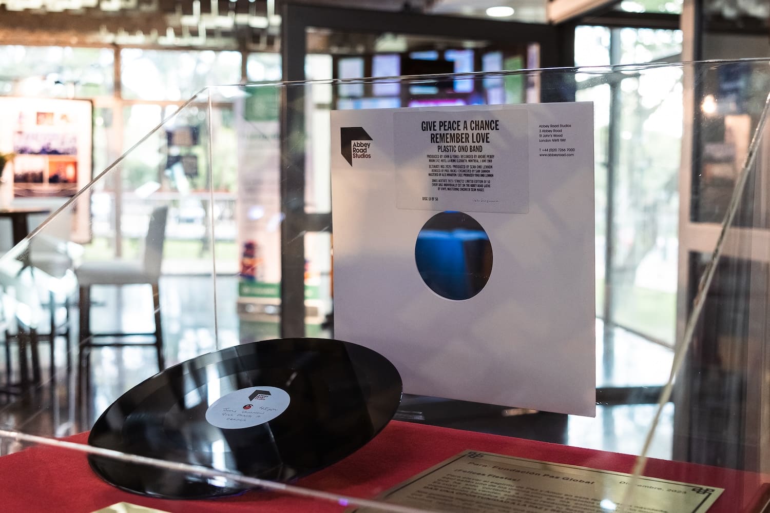 A vinyl record and sleeve sit elegantly in a glass case, set against the blurred interior of a building with tall windows. This display echoes the harmonious efforts of global peacebuilding initiatives championed by organizations like the Global Peace Foundation in Paraguay.