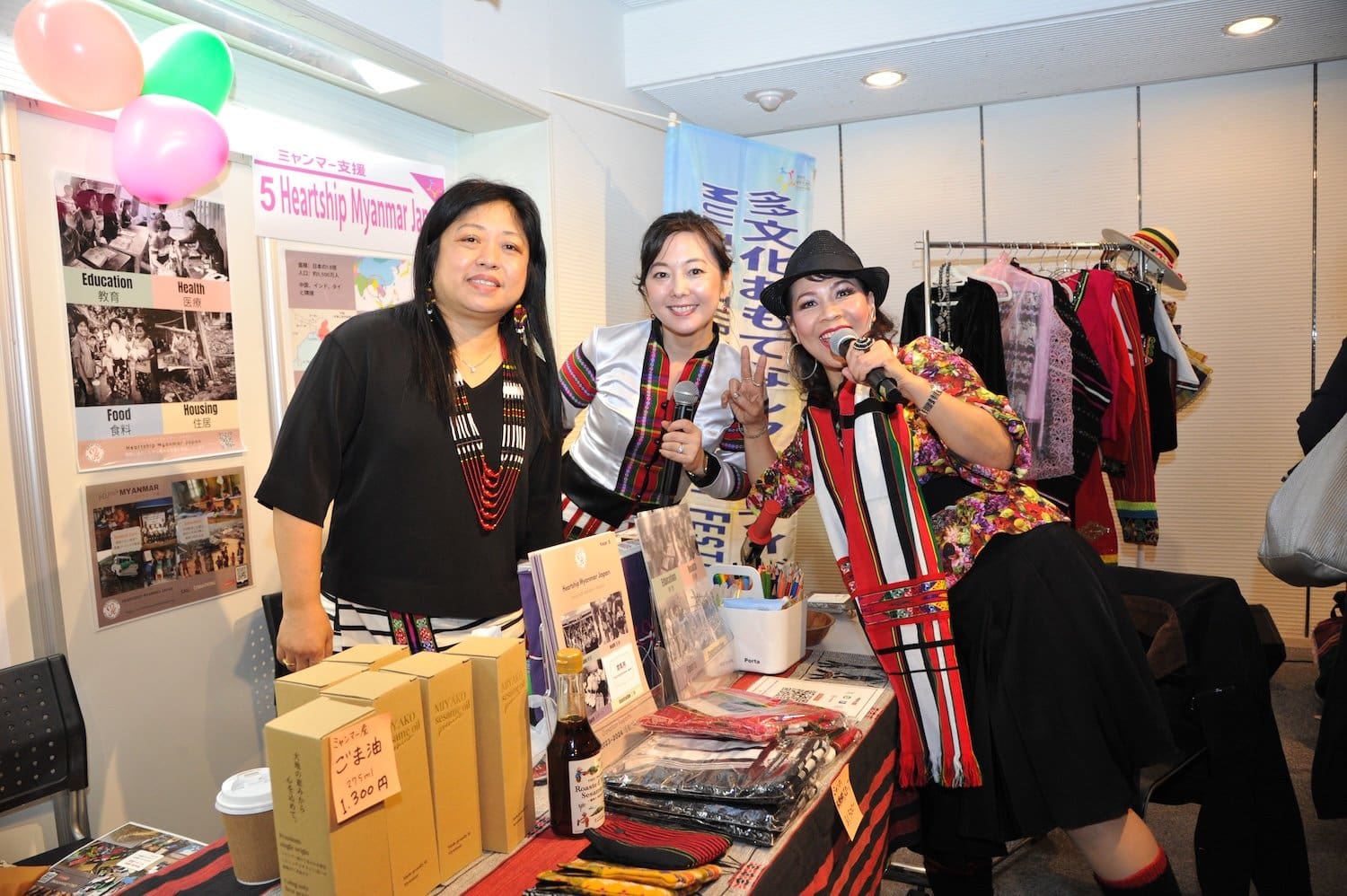 Three women in traditional clothing stand at a booth, their joyful spirits connecting smiles as they showcase cultural items and share insights. It’s a vibrant scene at this multicultural family festival, celebrating diversity and community.