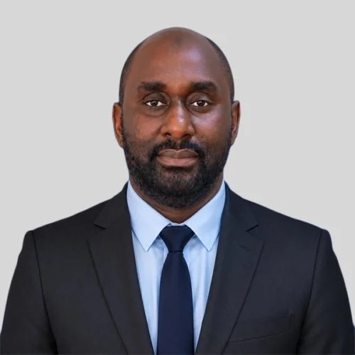 Hussein Sengu, dressed in a black suit, light blue shirt, and navy tie, poses against a plain light gray background.