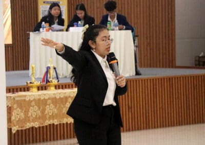 A woman in a suit speaks passionately into a microphone during the socio-debate, gesturing with her arm. In the background, a panel of three seated individuals with papers and bottles is visible, setting the stage for this pivotal 2024 event in Cambodia.