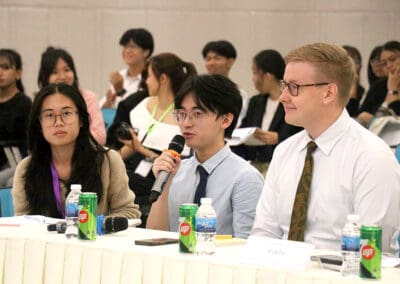 Three people sit at a conference table marked "JUDGES," one gripping a microphone. The table is adorned with beverages, notebooks, and nameplates. In the background, audience members eagerly await insights from the 2024 Socio-Debate focused on Cambodia's dynamic future.