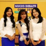 Three young women in matching white tops and blue skirts stand confidently in front of a "Socio Debate Open 2022" sign, showcasing their camaraderie. The anticipation for the Socio-Debate event in Cambodia 2024 is palpable as they prepare to bring their voices to the global stage.