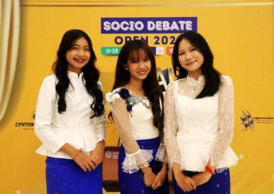 Three young women in matching white tops and blue skirts stand confidently in front of a "Socio Debate Open 2022" sign, showcasing their camaraderie. The anticipation for the Socio-Debate event in Cambodia 2024 is palpable as they prepare to bring their voices to the global stage.