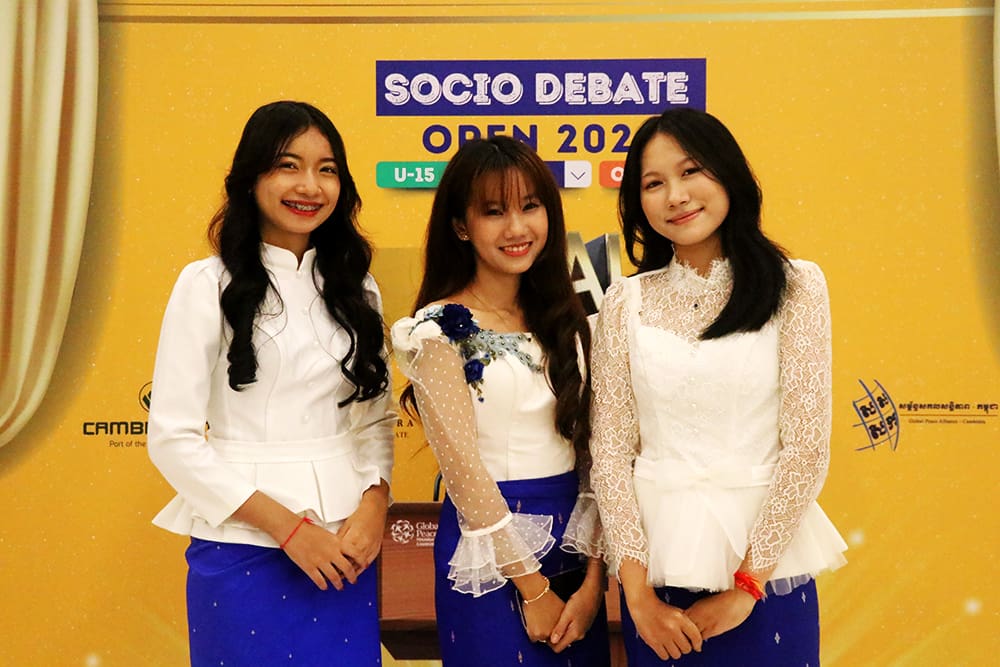 Three young women in matching white tops and blue skirts stand confidently in front of a 