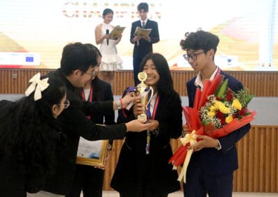 A group of people celebrate on stage, presenting a trophy and flowers at the 2024 Socio-Debate event in Cambodia.
