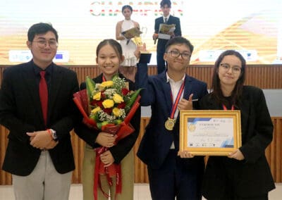 Four people stand on stage at the 2024 Socio-Debate in Cambodia; one holds flowers, another proudly displays a certificate and medal. They are elegantly dressed in formal attire, capturing the moment's significance with grace and poise.