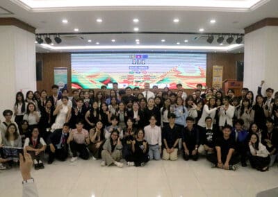 A large group of people posing together indoors in front of a colorful screen displaying graphics for the Socio-Debate 2024 event.