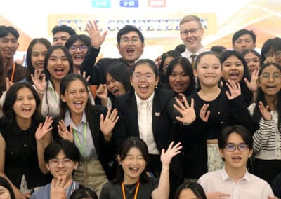 A diverse group of people, dressed formally, smiling and waving at a camera indoors, as they gather for a vibrant socio-debate on Cambodia's 2024 developments.