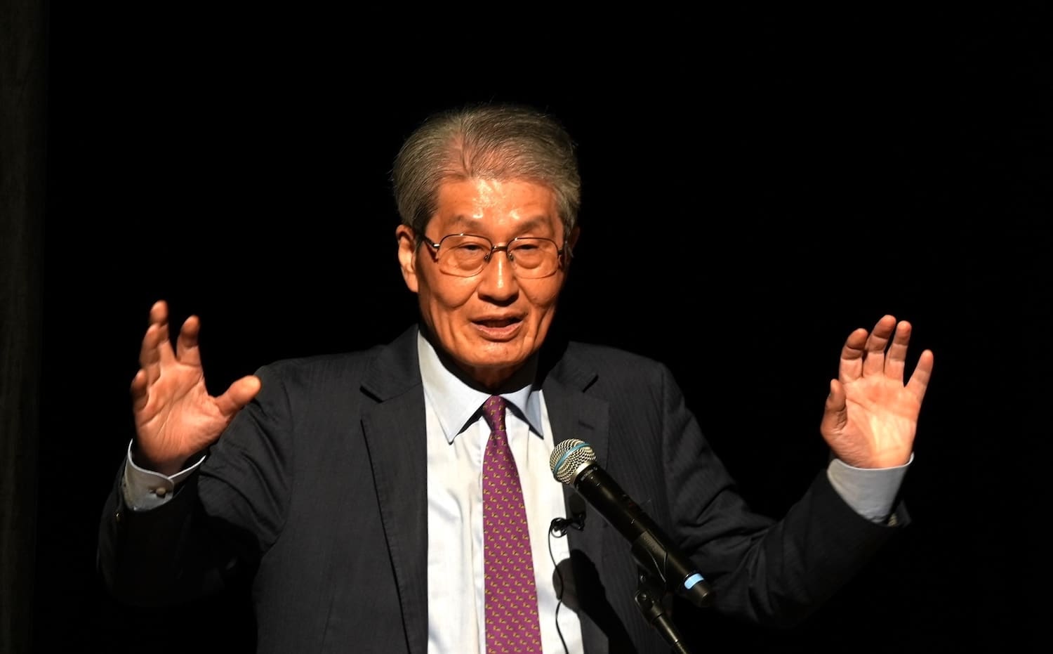 At the Peace Design Forum, an elderly man in a suit stands on stage speaking into a microphone with his hands raised, passionately discussing peacebuilding efforts in Northeast Asia.