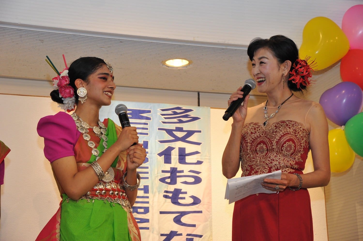 Two women on stage smile while speaking into microphones at a vibrant 2024 multicultural event. One wears traditional attire with vivid colors, the other dazzles in a red lace dress. Balloons float joyfully in the background, capturing the festive and inclusive spirit of this family festival.