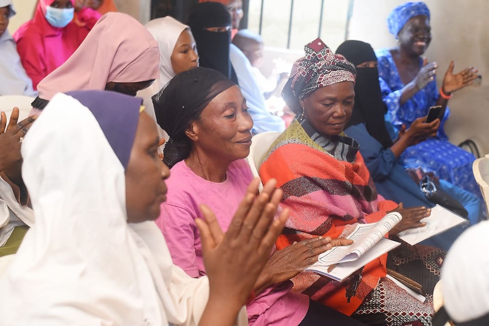 A group of women in Kaduna State, seated indoors and wearing colorful clothing and headscarves, engage in peacebuilding activities, some clapping and holding papers as they collaborate harmoniously.