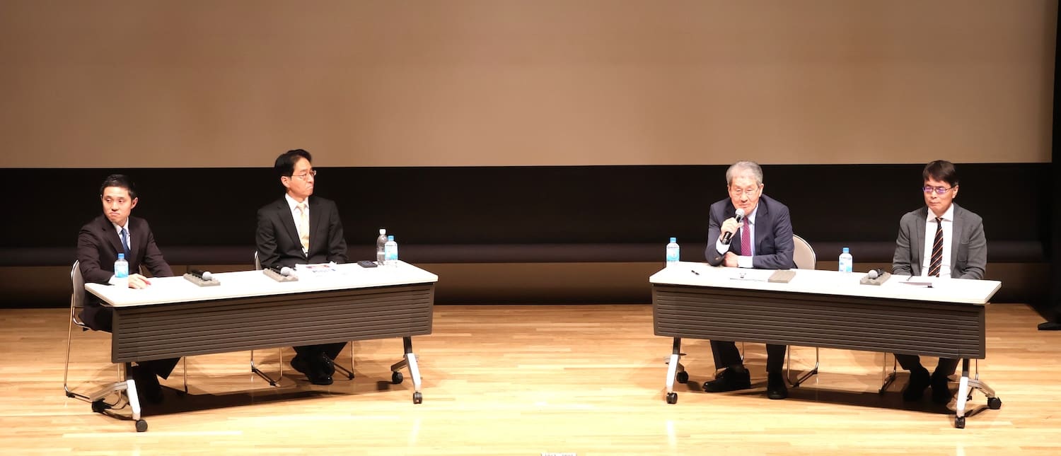 At the Peace Design Forum, four men in suits sit at two tables on a stage with microphones and bottled water, engaging in discussions pivotal to peacebuilding in Northeast Asia.