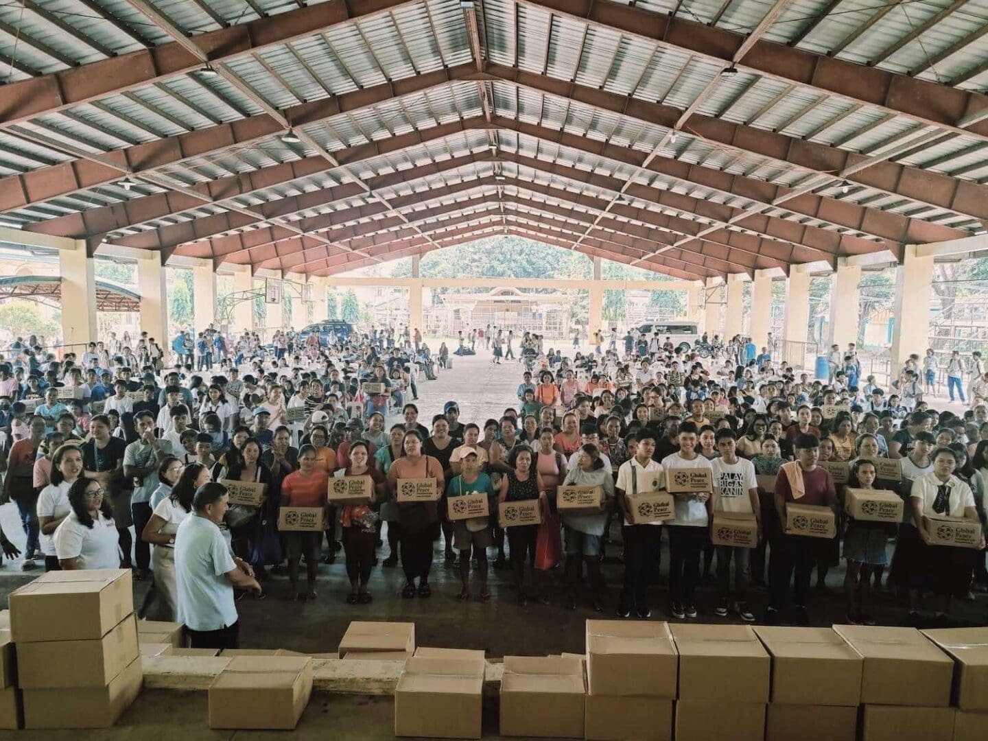 A large group of people stands in a covered area, holding boxes labeled 