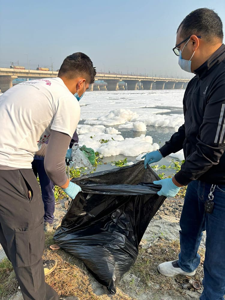 Amid a cleanup drive along the Yamuna Ghat, two individuals, donning gloves and masks, diligently collect trash in a large black bag by the foamy riverbank.