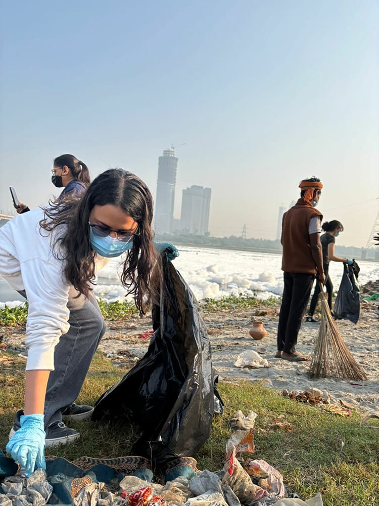 People wearing masks and gloves participate in a cleanup drive, picking up trash from a grassy area near Yamuna Ghat, with city buildings visible in the background.