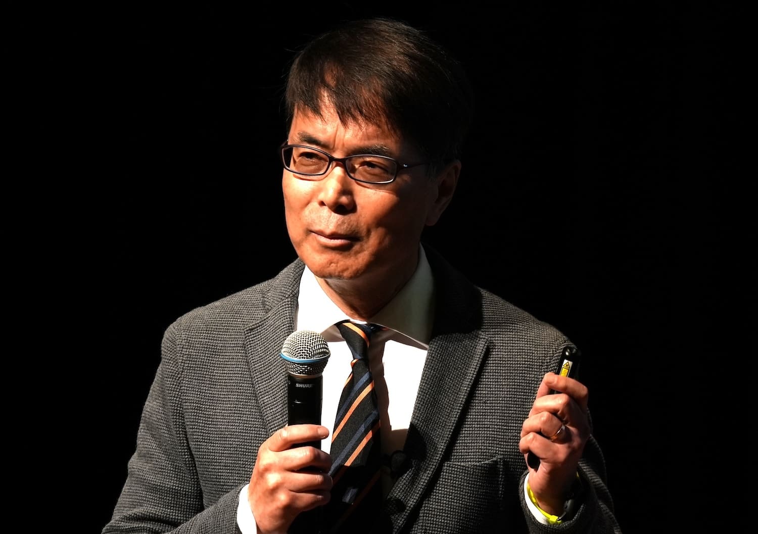 A man in a suit and tie holds a microphone and a small object, speaking passionately on stage at the Peace Design Forum. The dark background highlights his dedication to peacebuilding initiatives in Northeast Asia.
