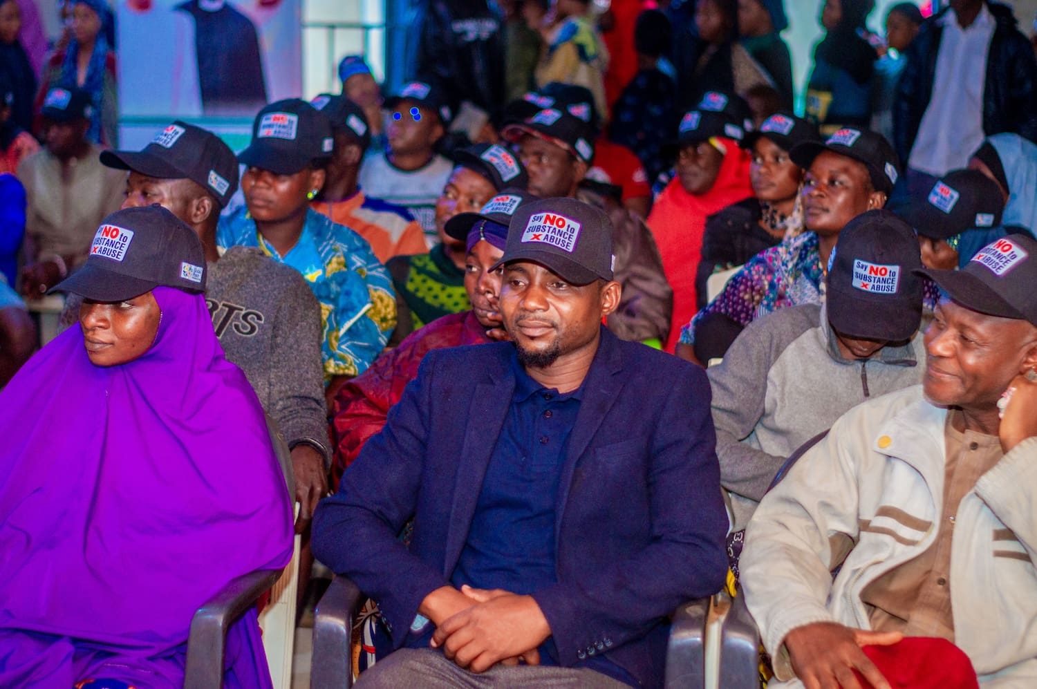 A group of people seated in a room, wearing matching black caps with a slogan from the Global Peace Foundation, highlighting their commitment to combating drug abuse in the Kaduna Community.