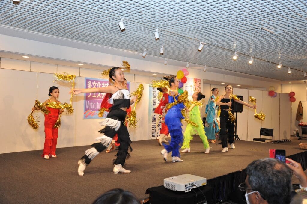 At a lively Family Festival in 2024, a group of six children in colorful outfits performs an energetic dance on stage. The beautifully decorated room buzzes with excitement as the multicultural audience cheers them on.