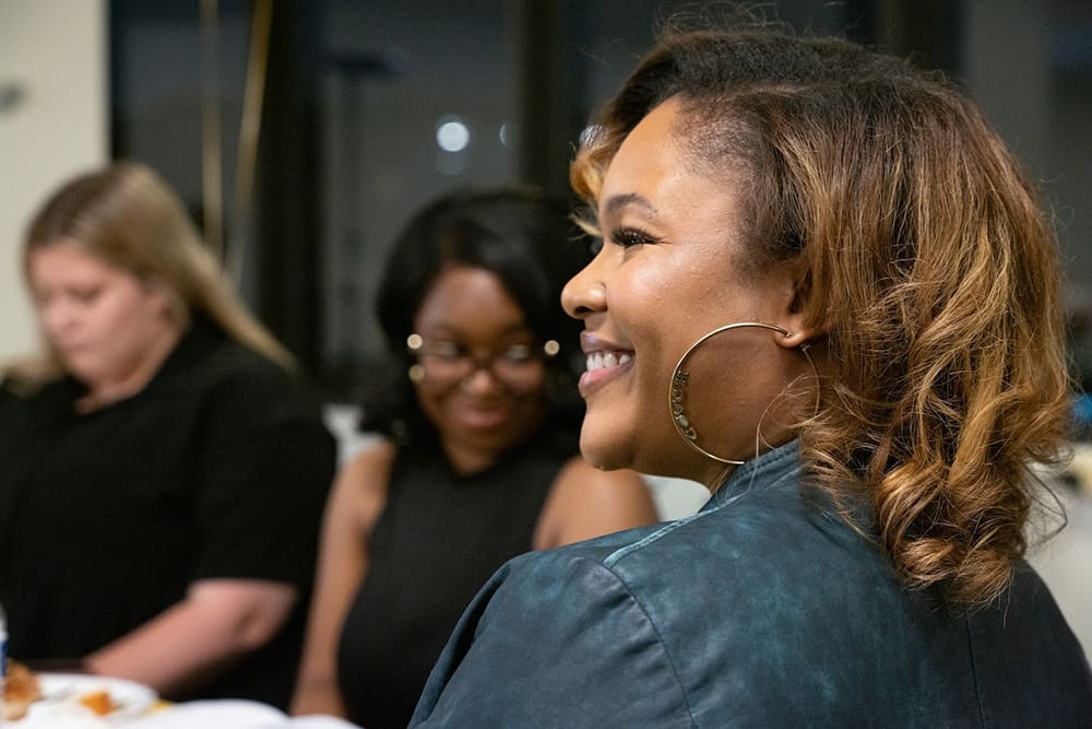 In a room filled with hope, a woman sporting hoop earrings smiles warmly while sitting beside two others, as if promoting peacebuilding initiatives in the heart of Baltimore.