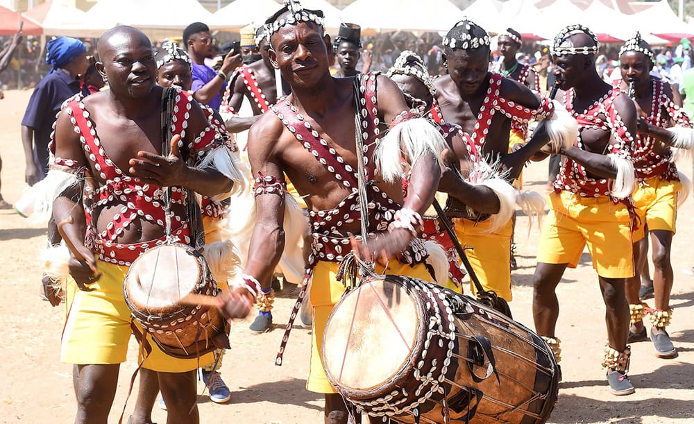 At the AFAN Festival 2025, men in traditional attire play large drums at a vibrant outdoor cultural event. They wear decorated outfits with beads and fringe, embodying unity as some perform enthusiastically with eyes closed, lost in the rhythm of their shared culture.