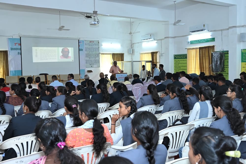 In a classroom setting, a speaker passionately addresses the audience about Education's role in shaping a Sustainable Future, with an insightful presentation projected on the screen. Attendees sit attentively on white chairs, eager to embrace the vision ahead.