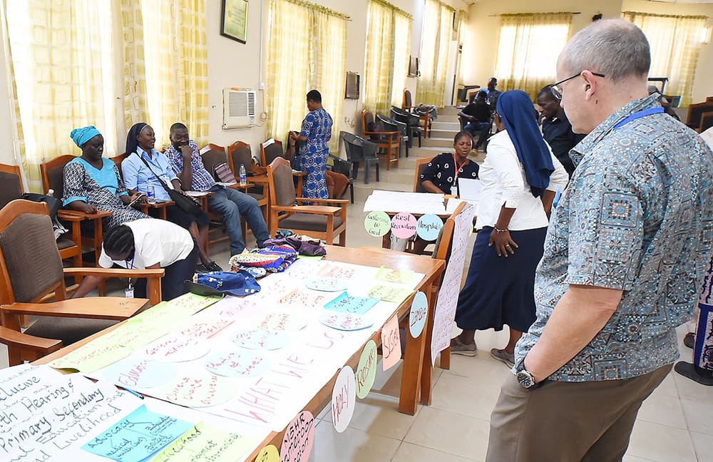 People gather in a room with tables displaying colorful posters and cloth items. Some are seated, others are standing and interacting.