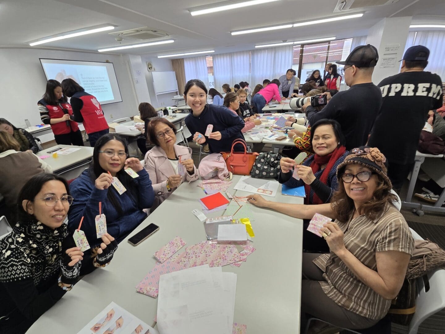 In a classroom, a group of people sits at tables, proudly displaying handmade crafts. As others work diligently in the background, the atmosphere buzzes with cross-cultural understanding and collaboration, advancing peacebuilding efforts and fostering leadership development between Japan and the Philippines.