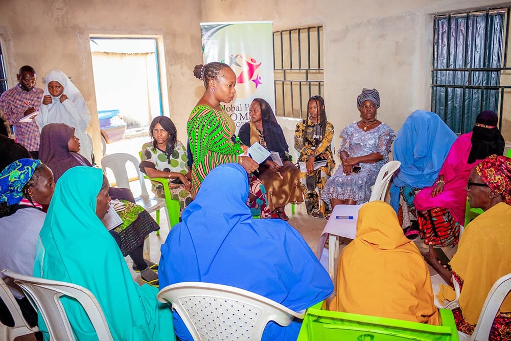 A group of people, mostly women in colorful attire, sit in a circle indoors for a meeting on women empowerment. A woman stands, passionately speaking to the group about financial literacy initiatives by GPF Nigeria.