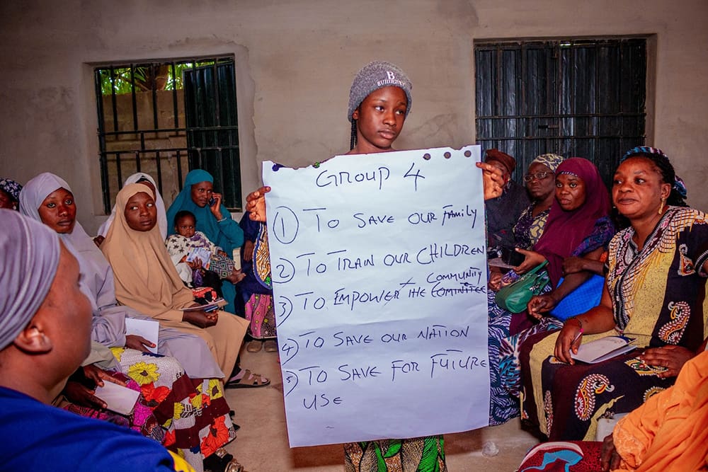 In a group discussion setting, a person holds a poster with handwritten goals on financial literacy training, surrounded by others seated. This initiative, supported by GPF Nigeria, emphasizes women empowerment through shared learning and growth.