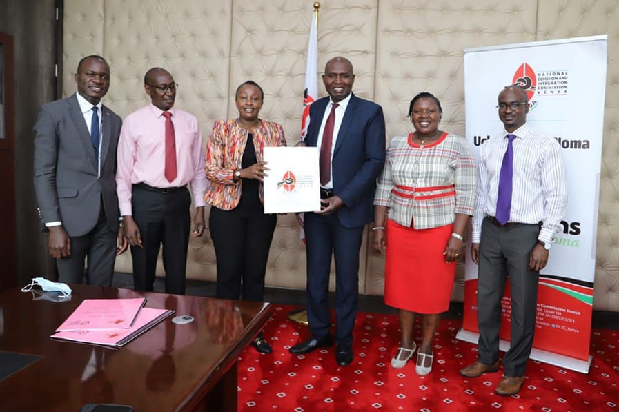 A group of six people stands together in an office, with one holding a folder. They pose for a photo next to a banner, proudly supporting the National Cohesion initiative to promote peace.