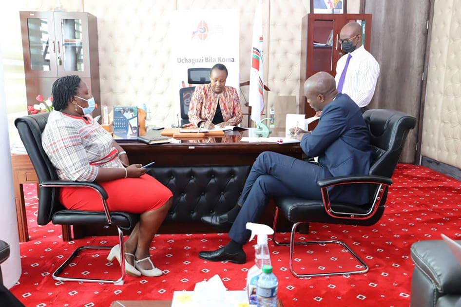 In an office setting, three people in formal attire are seated at a desk engaged in conversation while one stands by, embodying the spirit of the National Cohesion and Integration Commission's mission to promote peace and understanding.