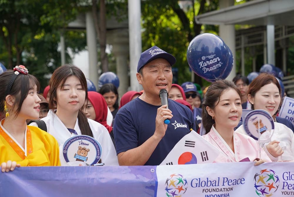 A group of people hold a banner at an outdoor event focused on peace. One person speaks into a microphone about harmony on the Korean Peninsula. Several participants, including youth, are wearing traditional Korean attire, adding cultural richness to the gathering.