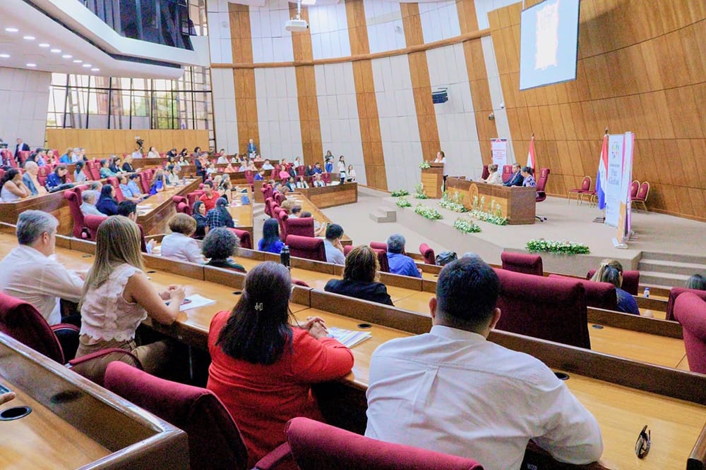 A large hall in Paraguay hosts a formal event where people seated in rows face a podium and screen, participating in discussions on empowering Indigenous Peoples for inclusive development.