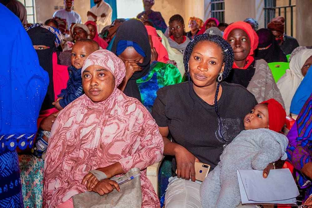 In a bright, crowded room, women and children gather indoors, some holding babies. A woman with a mobile phone and a sheet of paper exemplifies women empowerment during this financial literacy training organized by GPF Nigeria.