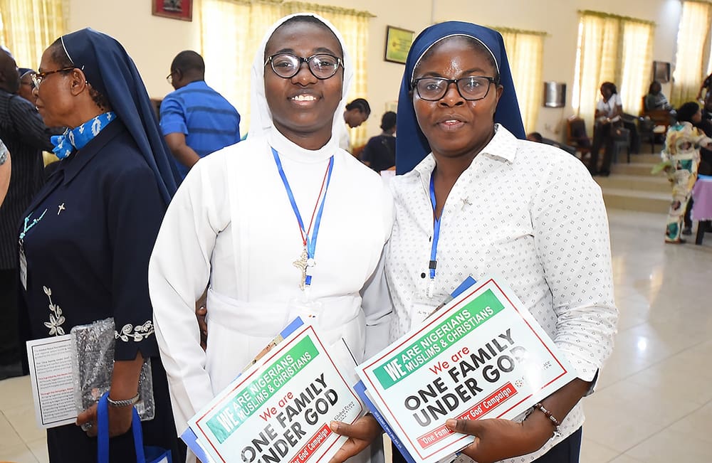 Two women wearing glasses and headscarves, one in a white habit, hold documents titled 