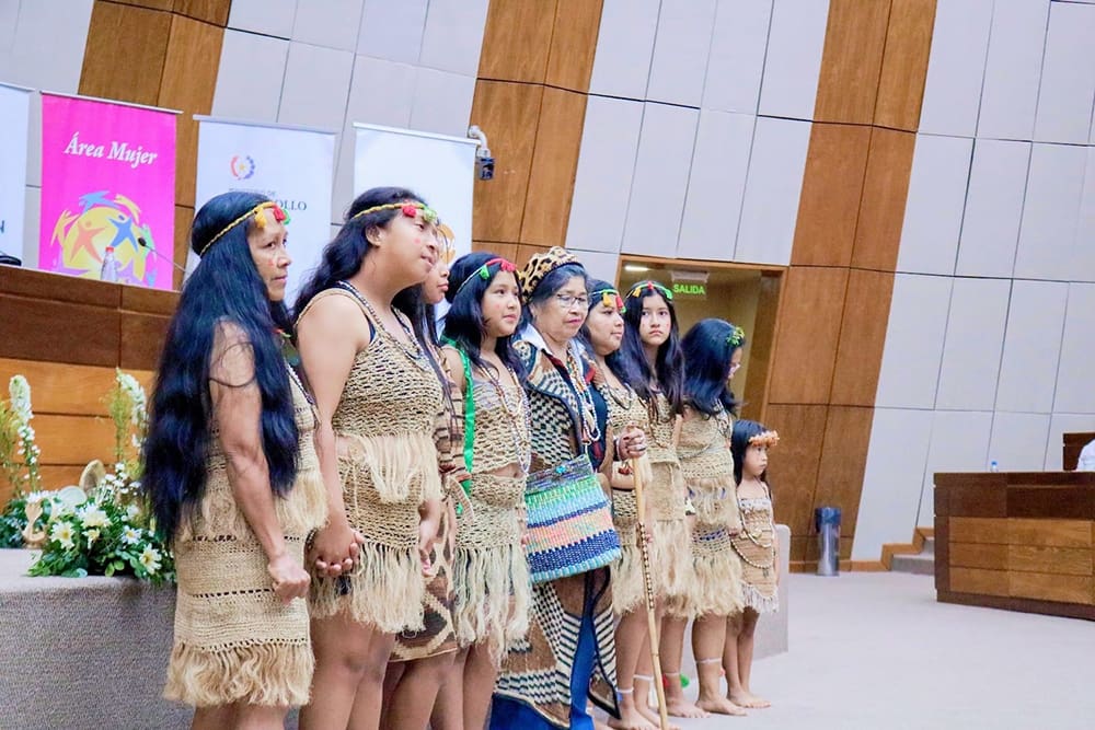 A group of women and girls in traditional woven clothing stand together in a modern hall with wooden paneling, next to a display and floral arrangements, representing the empowering indigenous peoples of Paraguay.