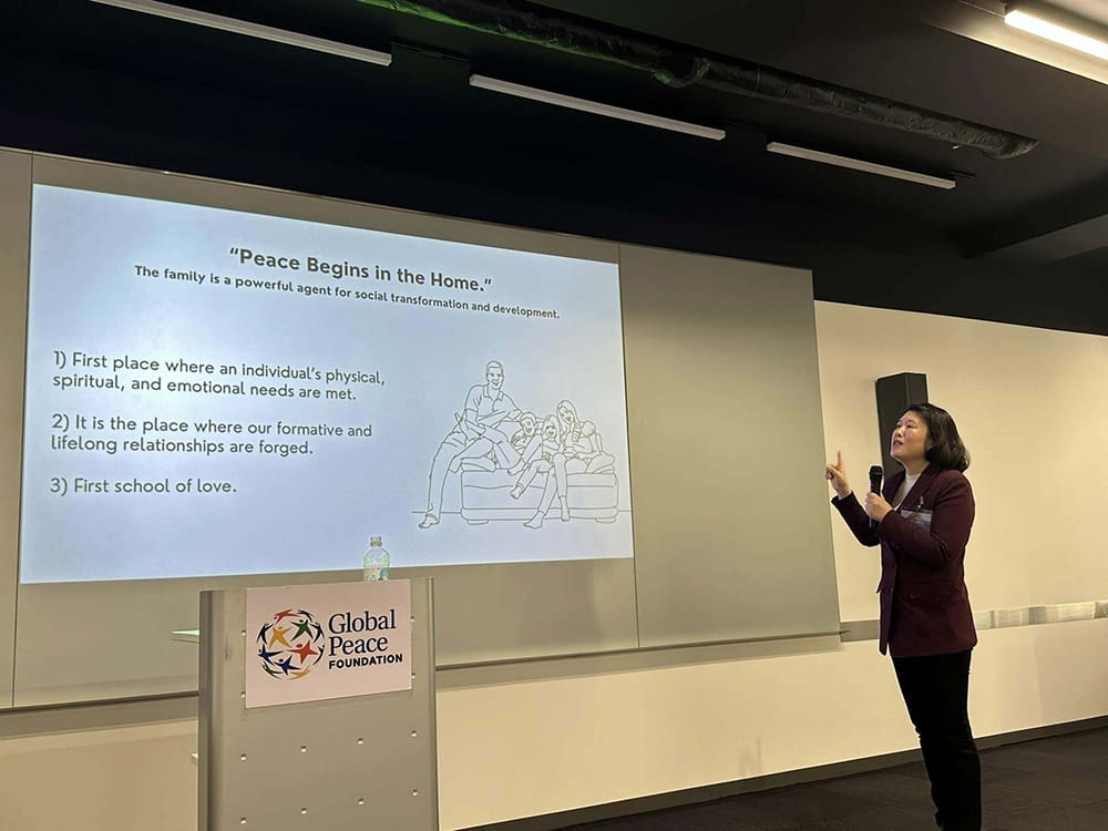 A person stands, gesturing at a screen with a slide titled "Peace Begins in the Home" during a Global Peace Leadership Exchange event in the Philippines.