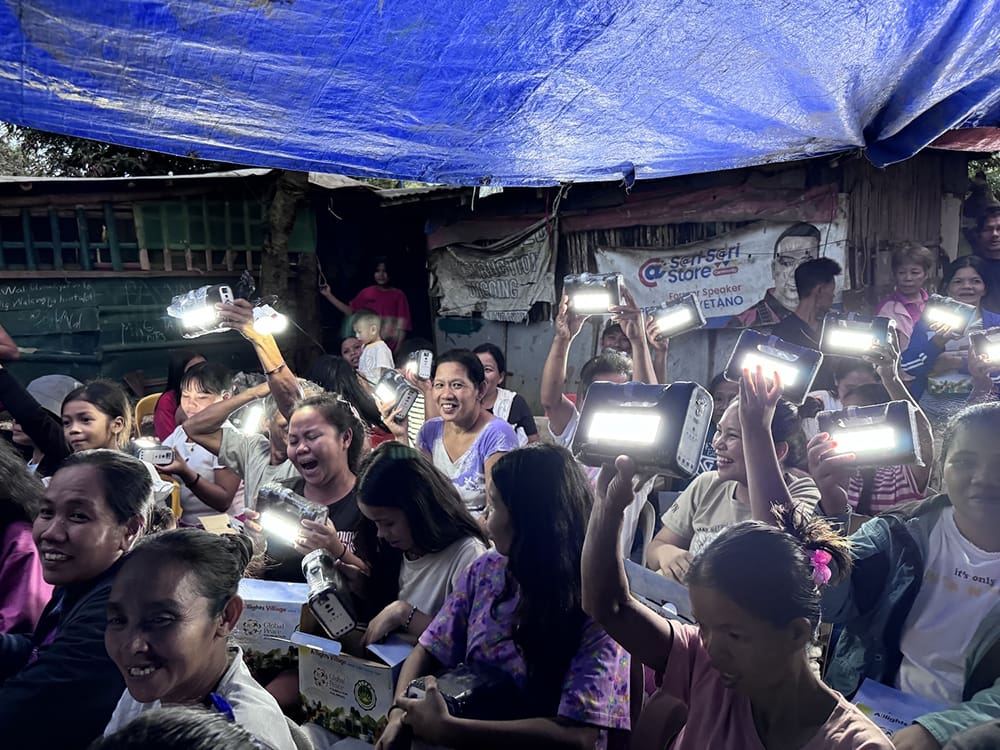 A group of people in Candelaria sits under a tarp, smiling as they hold up solar lanterns provided by the Global Peace Foundation. These small LED lights illuminate their gathering, symbolizing hope and unity for families across the Philippines.