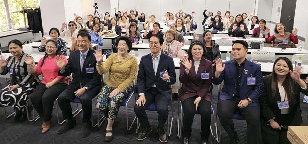 A group of people sitting in rows in a conference room, waving and smiling towards the camera, at the Global Peace Leadership Exchange event. Attendees from diverse backgrounds, including Japan and the Philippines, come together to foster understanding and collaboration.