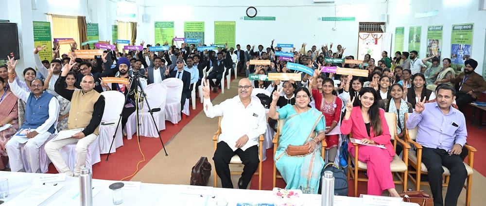 A group of people sitting in a large room, holding colorful signs and smiling at the camera. Some are seated in the front row and others are in chairs at tables.