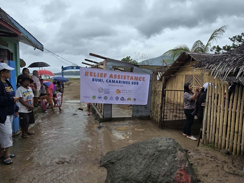 People standing in a muddy area near a banner reading "Relief Assistance Buhi, Camarines Sur" under a cloudy sky, as part of the GPF Philippines' efforts to rebuild the community.