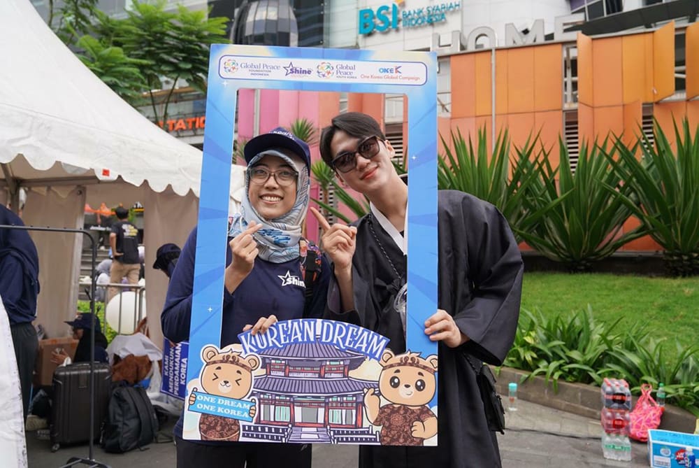 Two people pose with a "Korean Dream" photo frame at an outdoor event, capturing the spirit of peace on the Korean Peninsula. They smile and gesture with fingers, standing near tents and a building adorned with green plants in the background.