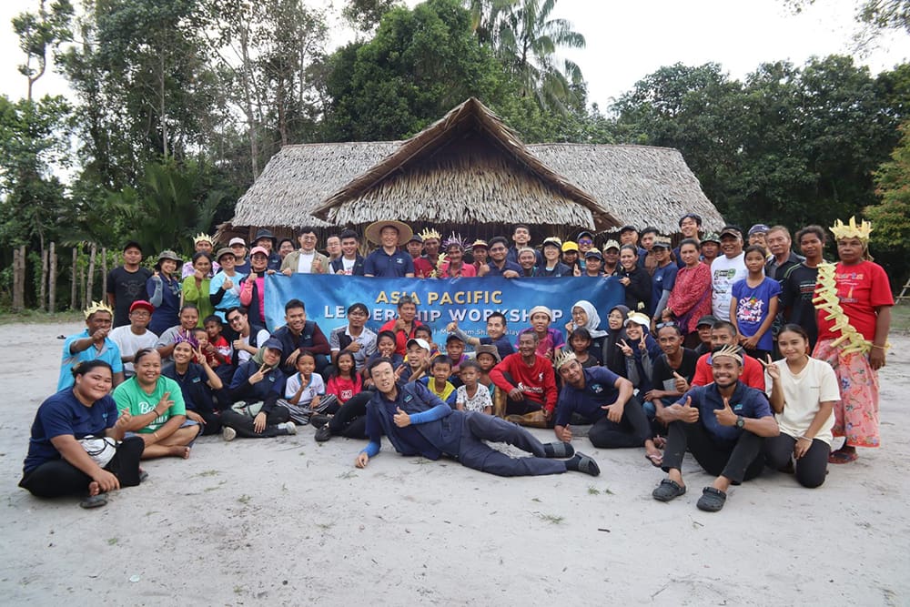 A large group of people poses in front of a thatched building, holding a banner reading "Asia Pacific Leadership Workshop." Trees are visible in the background.