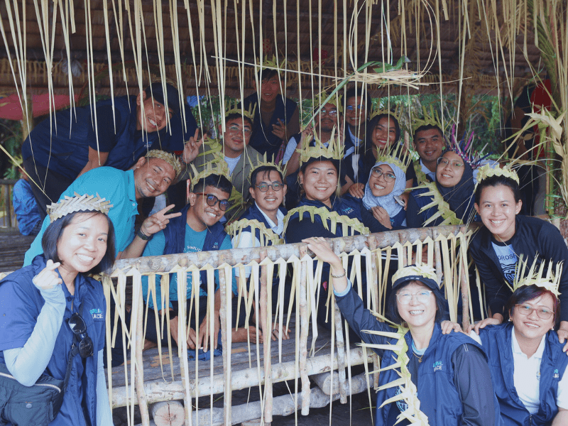 A group of people wearing woven leaf hats poses together inside a rustic wooden structure with more leaves hanging around.