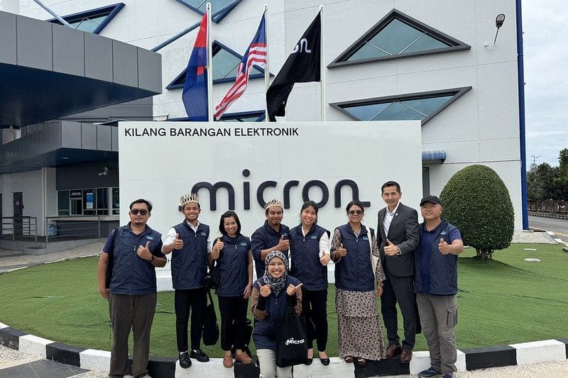 A group of people posing and giving thumbs up in front of a Micron sign at an electronics facility, celebrating the initiative where Micron and Global Peace partner to bring clean water to Orang Asli communities, with flags waving proudly in the background.