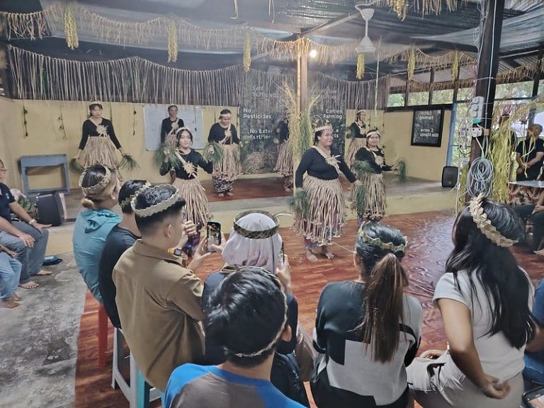 People wearing traditional attire perform a cultural dance inside a room with onlookers seated and watching.