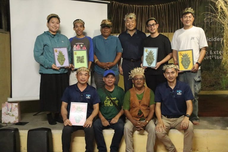 A group of ten people, some seated and others standing, pose with framed pineapple artwork and wear crown-like hats indoors.